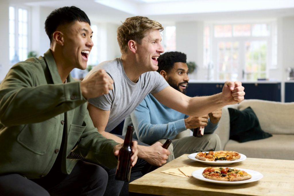 Group Of Excited Male Friends Watching Sports On TV At Home In Lounge With Pizza Together
